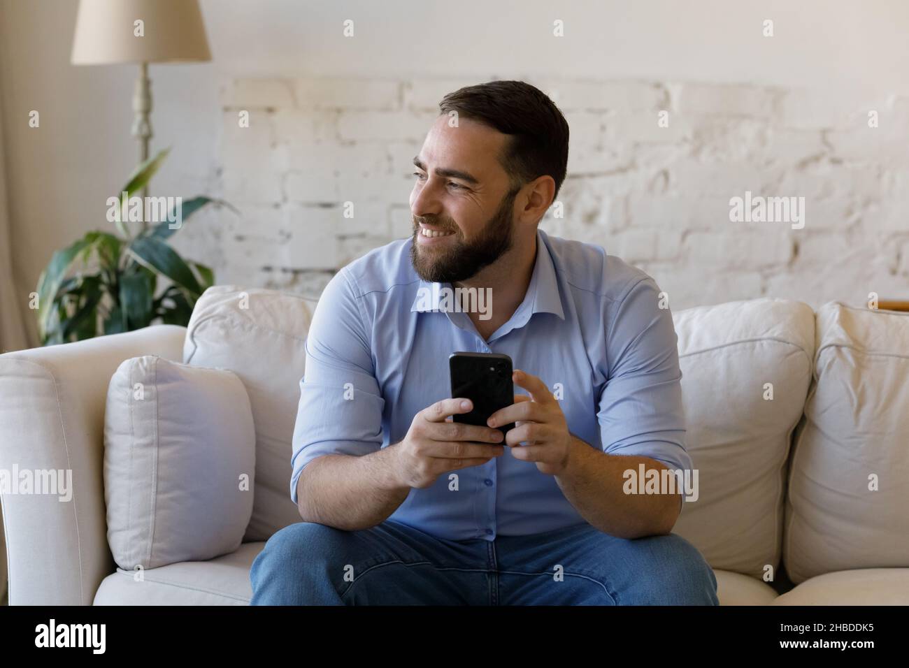 Smiling relaxed dreamy young man using smartphone at home. Stock Photo