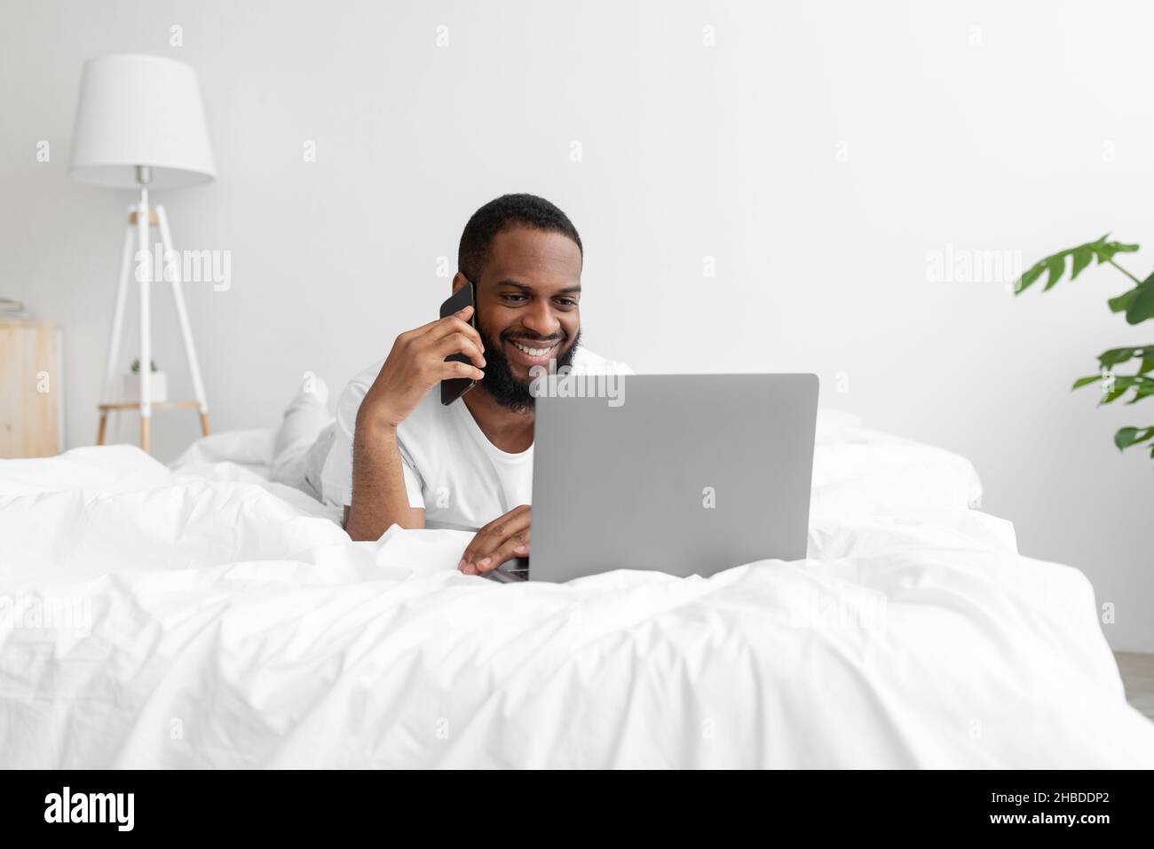 Smiling young black bearded male lies on bed in white minimalist bedroom interior, speaks by phone Stock Photo