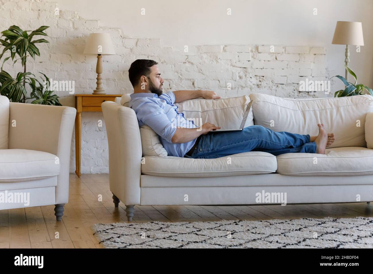 Concentrated young man using computer lying on couch. Stock Photo