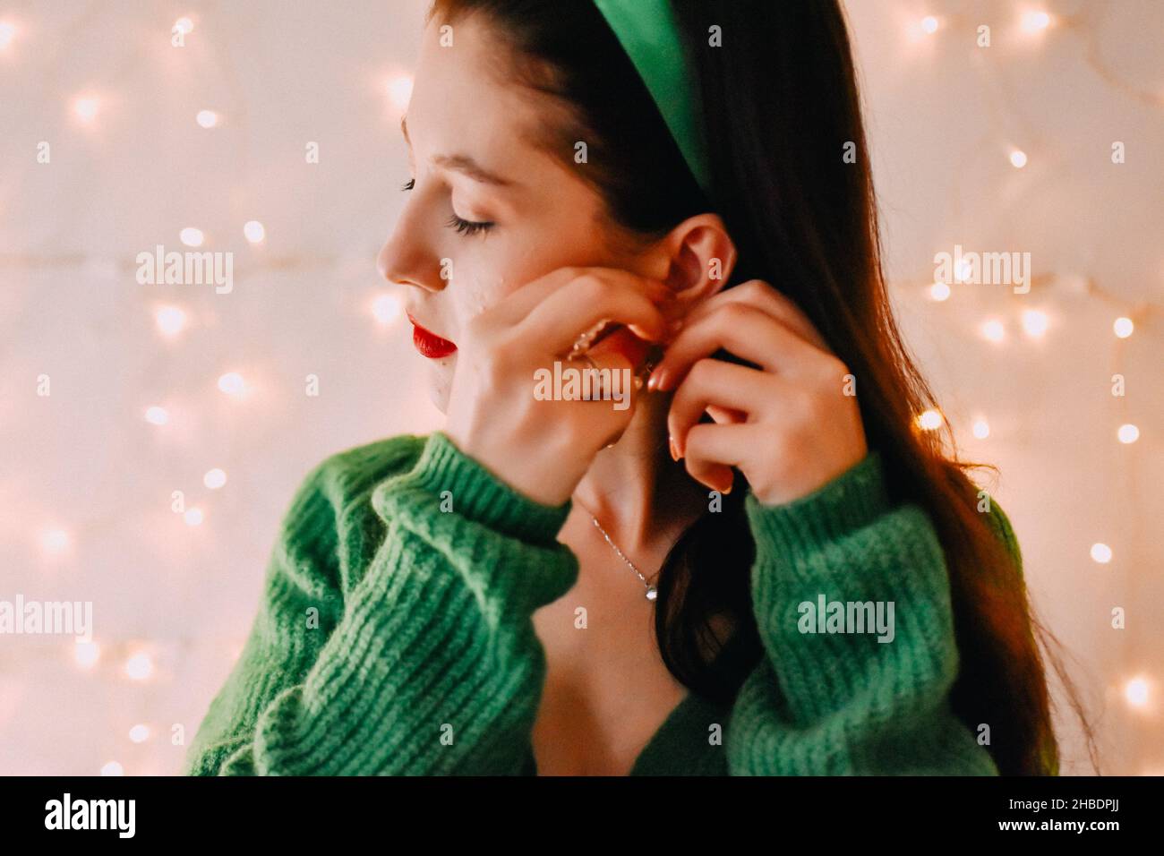 A closeup shot of a Caucasian girl putting on her earing and Christmas lights on the wall Stock Photo