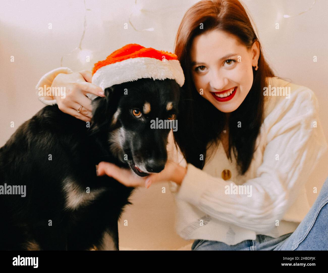 A closeup shot of a Caucasian girl smiling to the camera with her dog and Christmas lights on wall Stock Photo