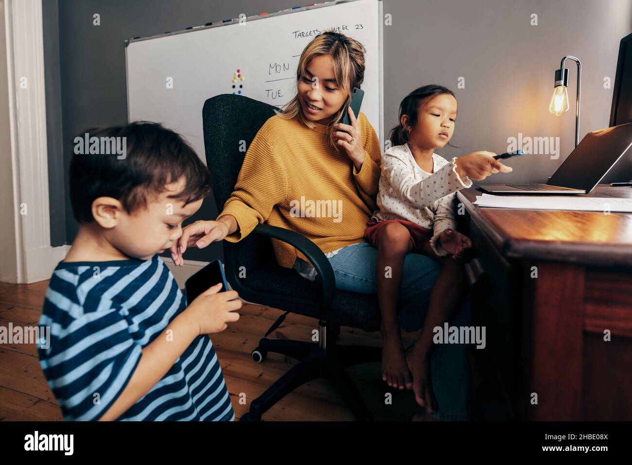 Multi-tasking mother of two working in her home office. Working mom taking a business phone call with her kids playing at her desk. Single mom communi Stock Photo