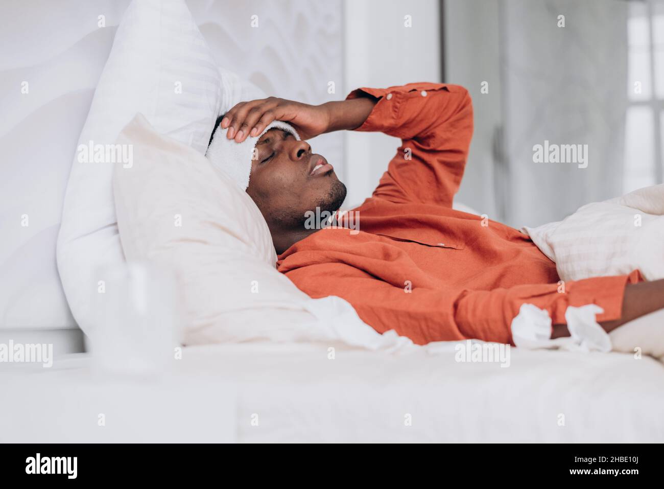 Young Afro-American man in orange shirt with patch on forehead suffers from high temperature resting in large bed close side view Stock Photo