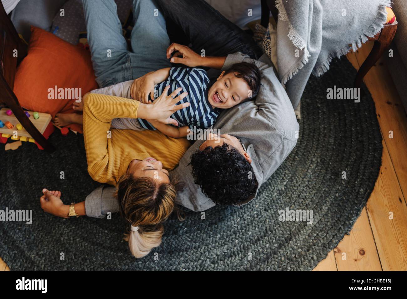 Overhead view of a little boy having fun with his mom and dad in their play area. Young boy laughing cheerfully while lying on top of his parents. Fam Stock Photo