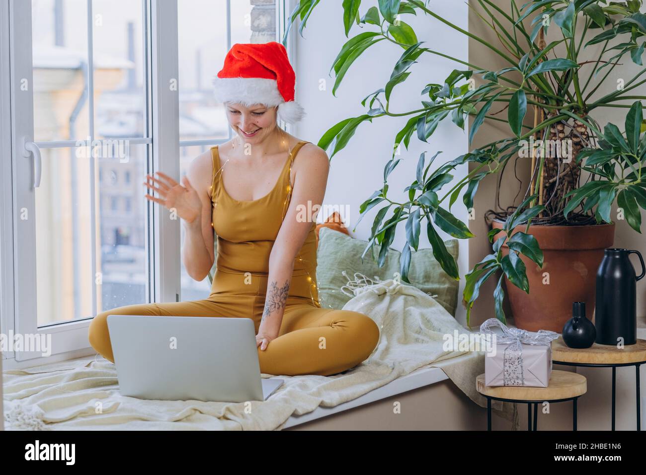 Young woman in Santa hat wrapped in illuminating garland talks via videocall with laptop sitting on blanket covering windowsill against plants Stock Photo