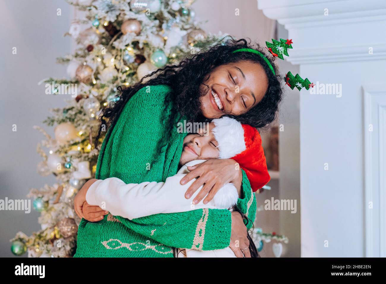 African american mom in green sweater and headband with christmas trees hugs her little daughter in a christmas hat against the background of a christmas tree. Stock Photo
