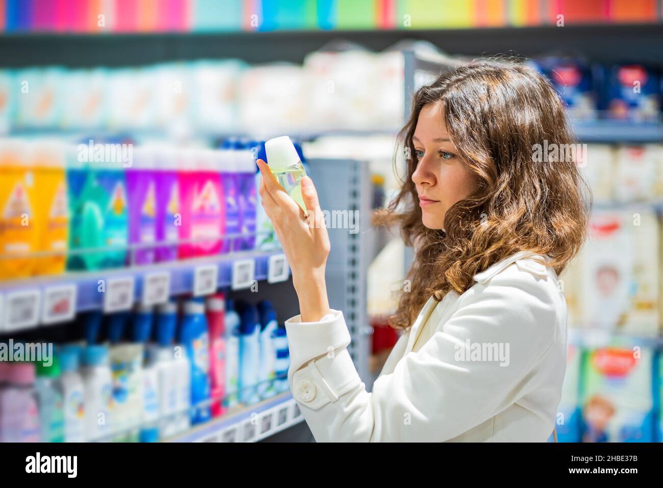 Brunette woman with long curly hair wearing white coat reads instruction on deodorant package standing in household goods store Stock Photo