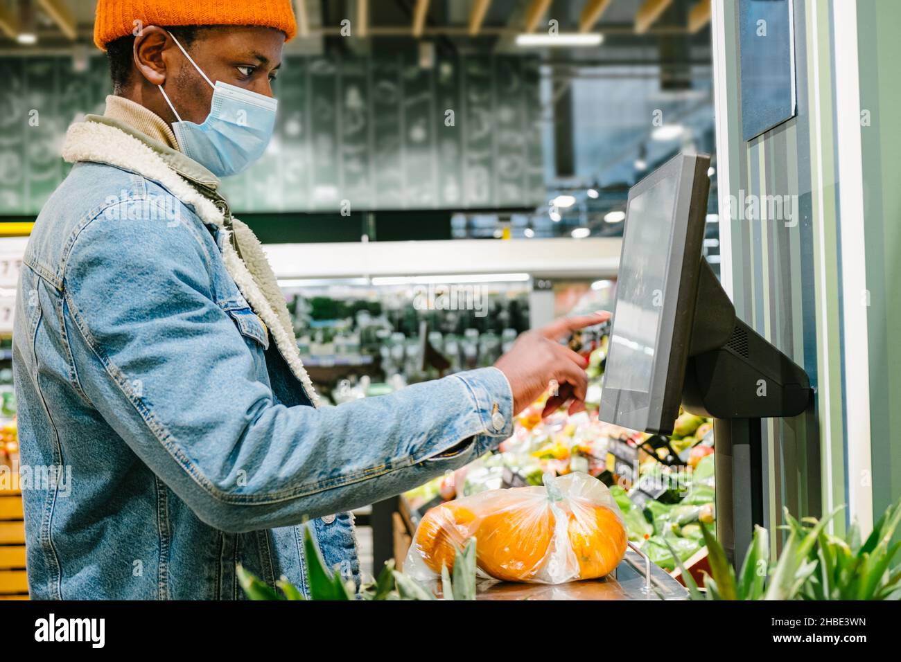 African hipster man in a medical mask weighs fresh oranges in a plastic bag on an electronic scale in a supermarket, selects the desired product on the touch screen Stock Photo
