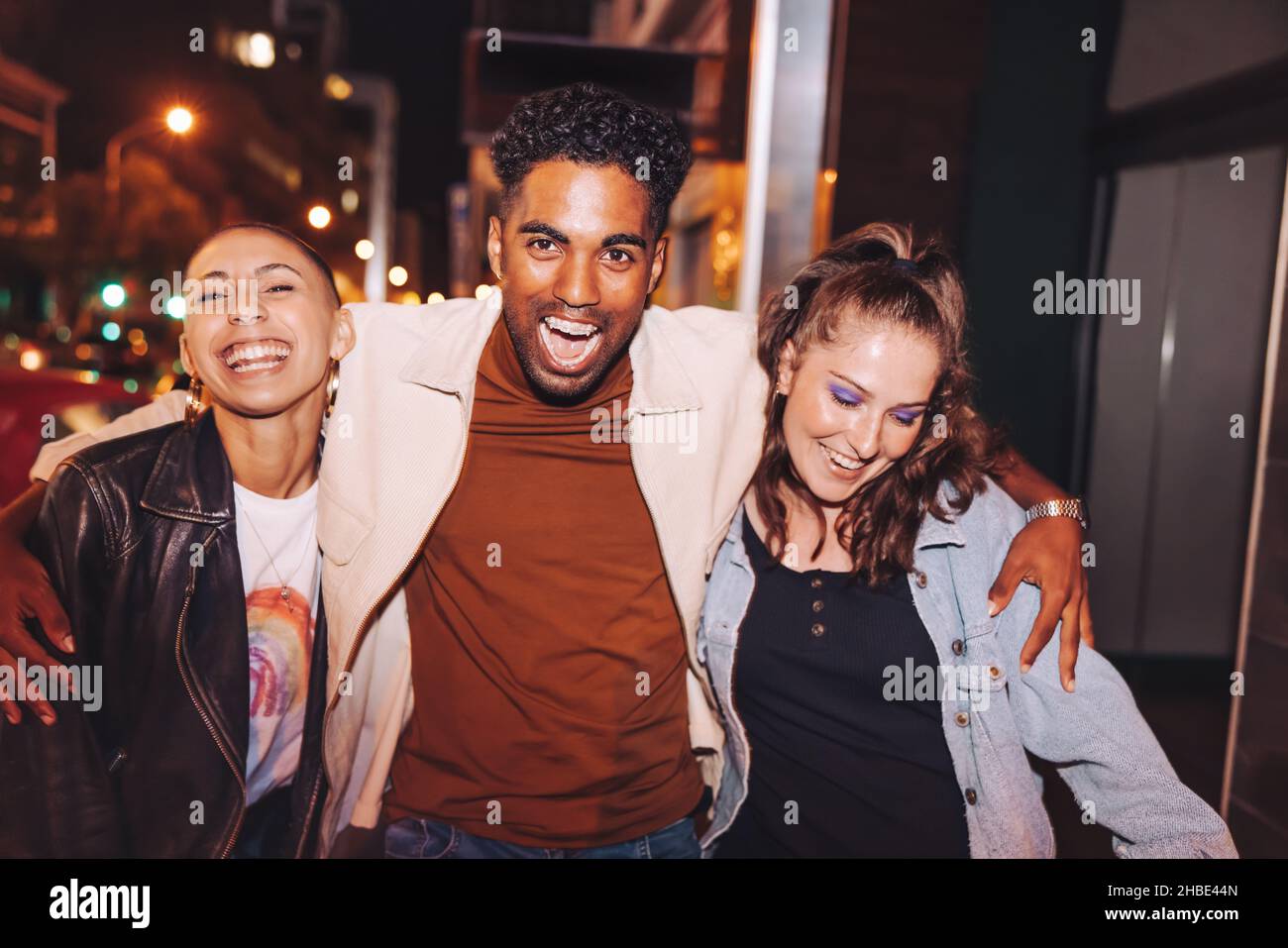 Three young friends having fun in the city at night. Group of vibrant young people laughing happily while walking together. Multicultural friends goin Stock Photo