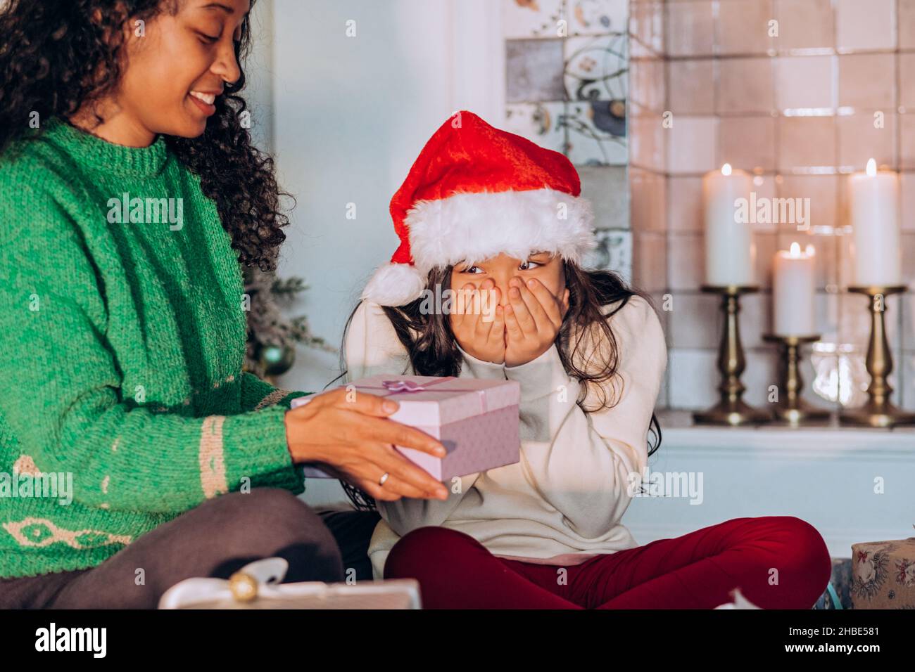 Curly African American woman and brunette long-haired daughter wearing holiday hats open present box sitting against Christmas tree Stock Photo