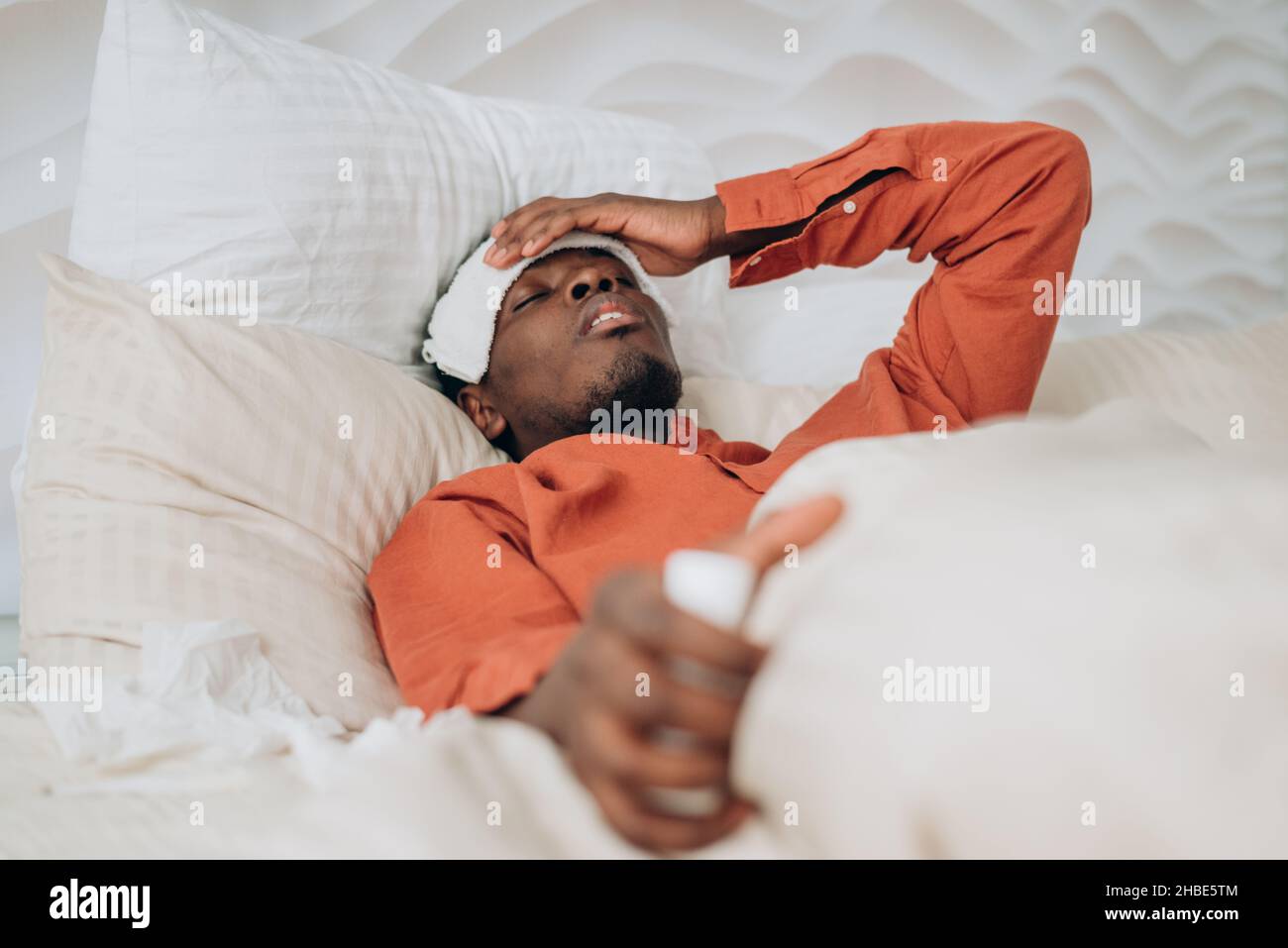 Upset African-American man in orange pajamas with patch on forehead suffers from cold lying in large comfortable bed close view Stock Photo