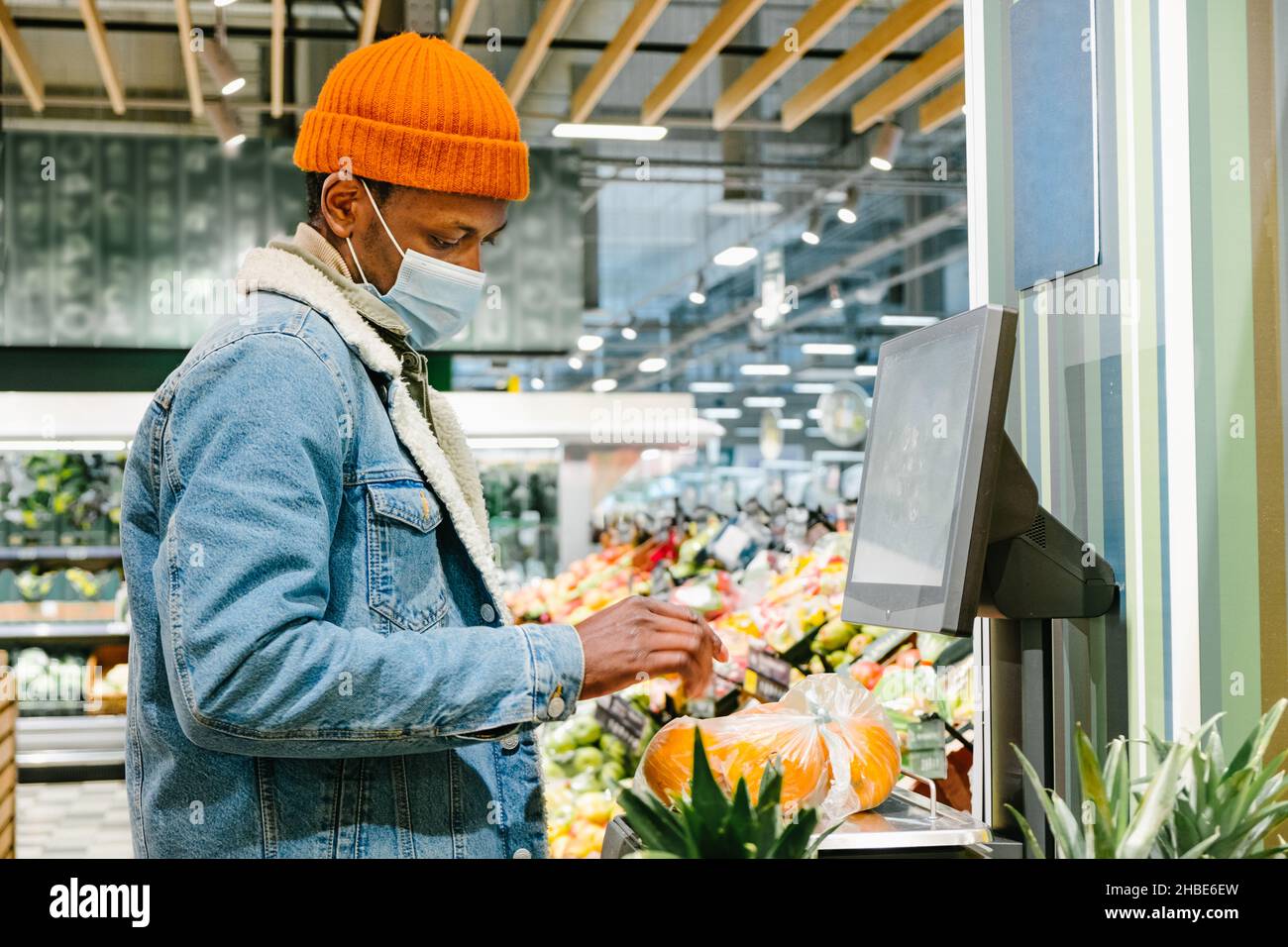 Stylish African-American guy in warm jacket with disposable mask weighs fresh oranges with digital scale in light supermarket Stock Photo