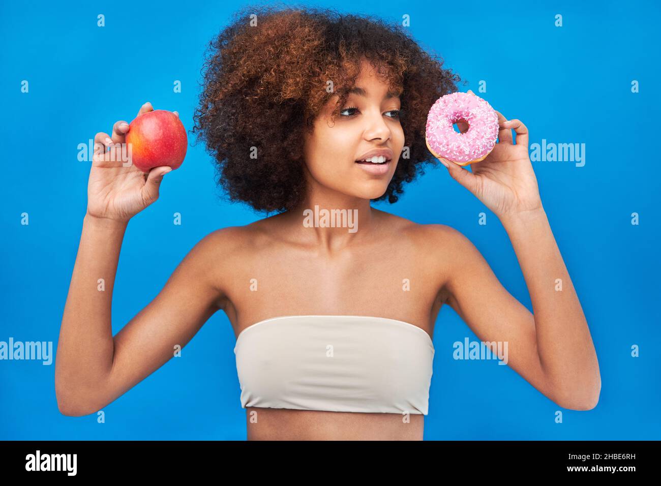 Curly haired mixed race young woman wearing light top poses holding apple and donut with frosting and smiles against bright blue wall Stock Photo