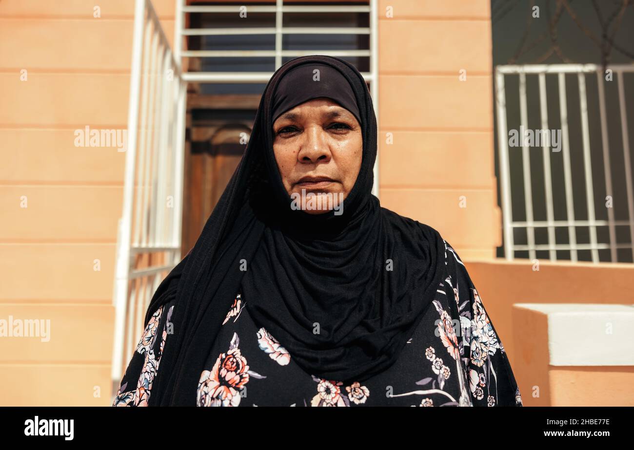 Middle-aged Muslim woman looking at the camera confidently while standing in front of her home during the day. Self-confident mid-adult woman wearing Stock Photo