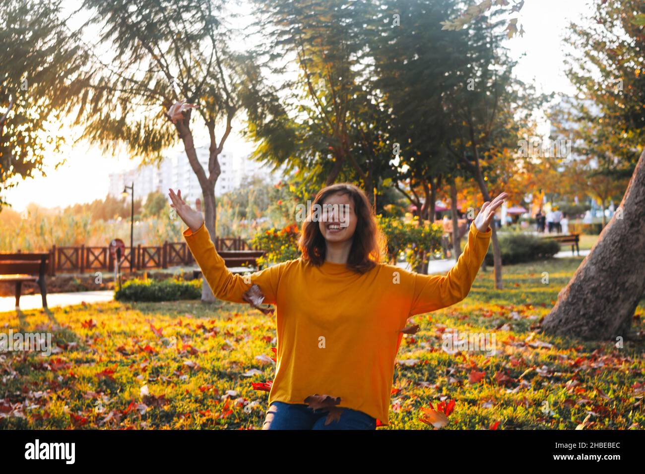 Curly young girl in yellow sweater and jeans sitting on grass and throws up dry leaves. Stock Photo