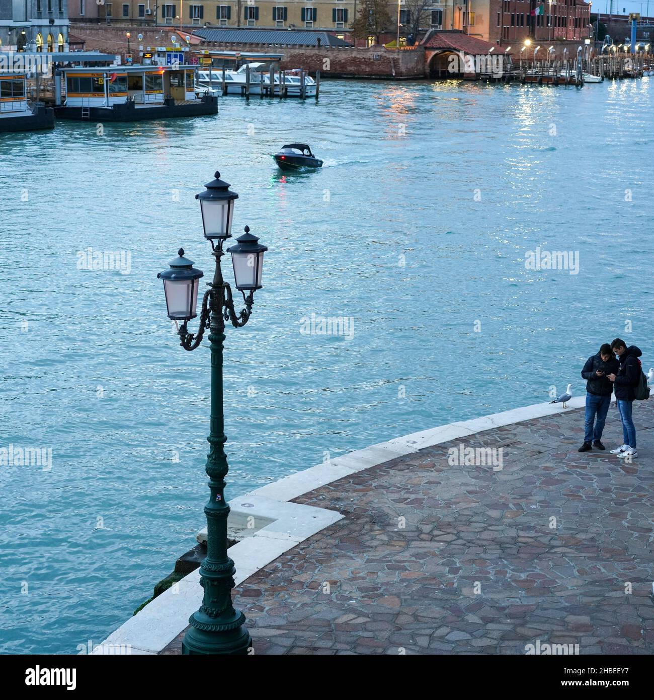 Two young men at a lamppost opposite the Piazzale Roma vaporetto water bus stop on the Grand Canal in Venice look at their smartphones. Stock Photo