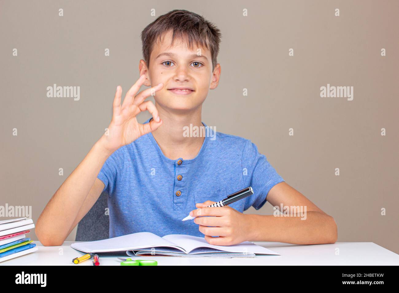 Boy hand holding pen in left hand and writing in a notebook, doing homework. Smiling happy kid enjoy learning and show OK hand gesture. Left Handers Stock Photo