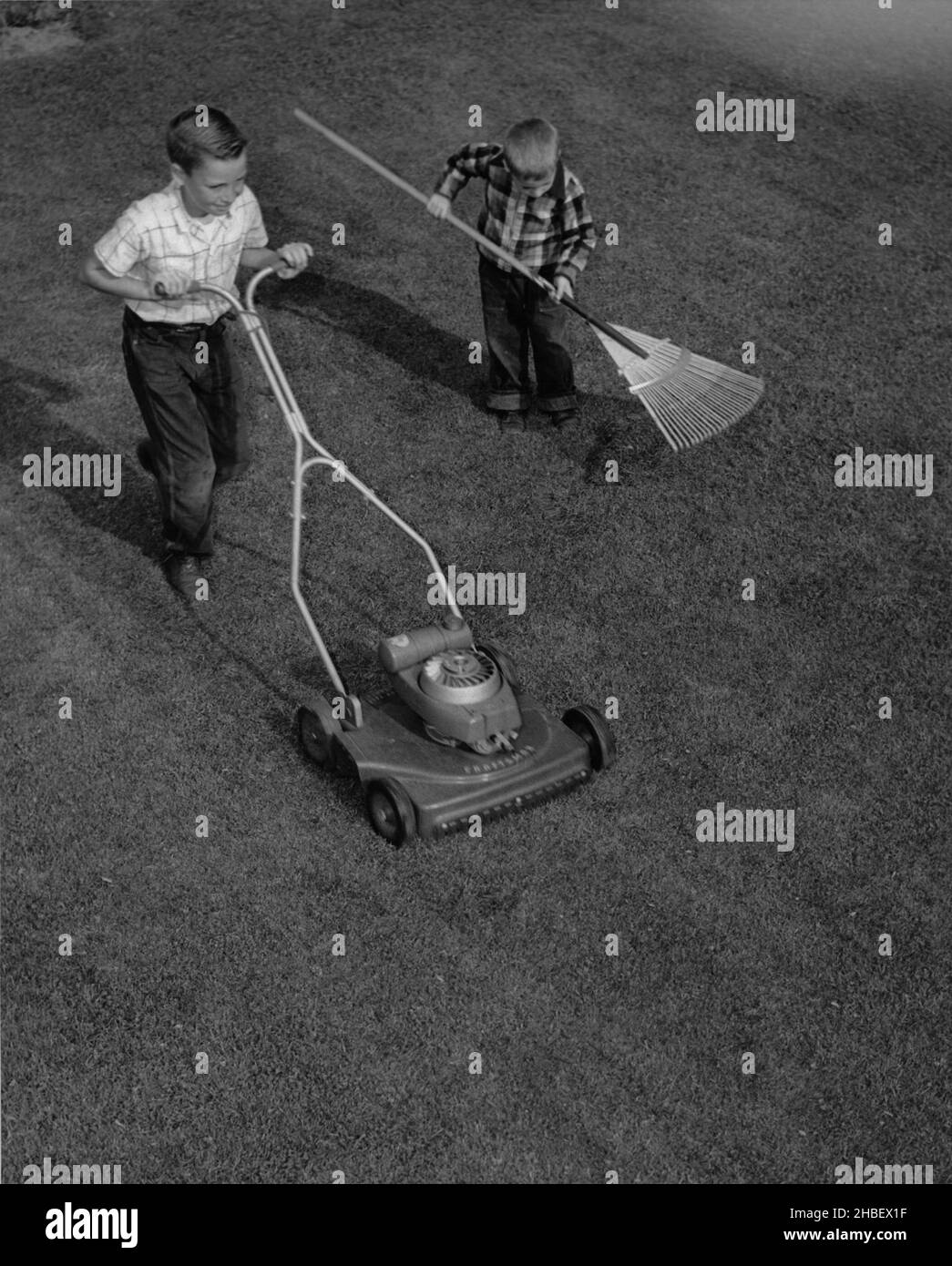 Two young boys doing yardwork. One pushing power mower and the other raking with leaf rake