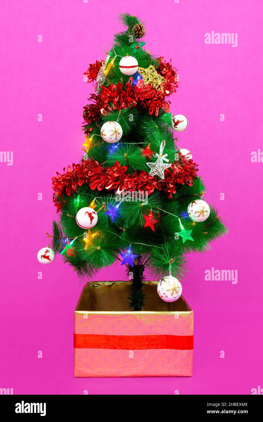 A small Christmas tree decorated with balls, stars, pine cones, garlands and colored lights. The base is inside a square box decorated with gold paper Stock Photo