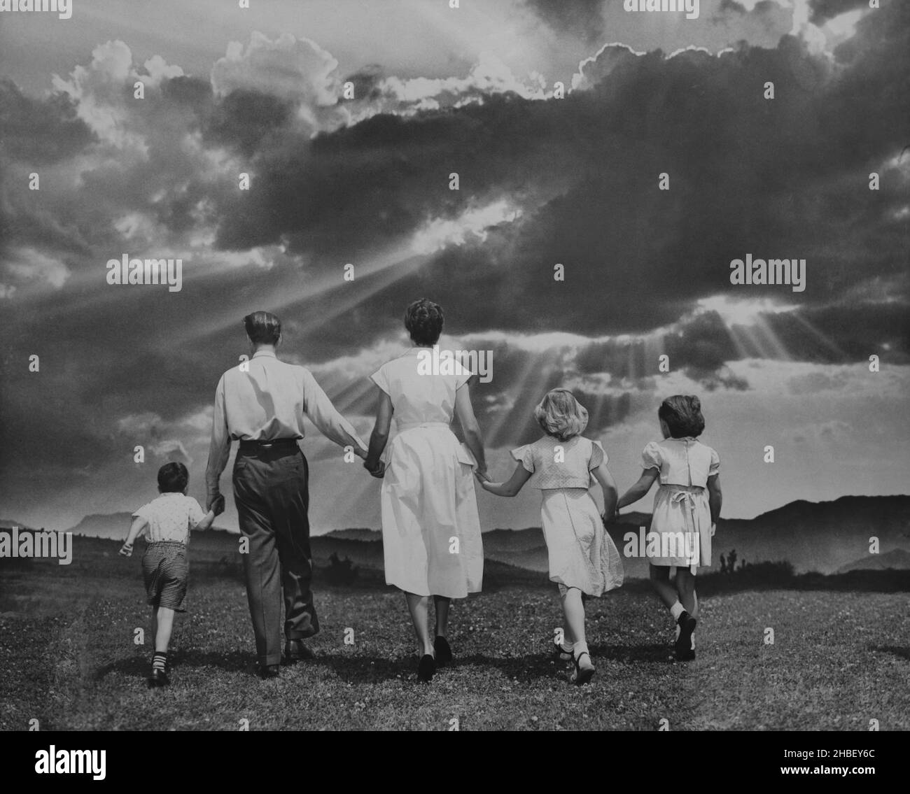 Mom, Dad and 3 children all holding hands walk along a grassy field into a sunburst. Circa 1960 Stock Photo