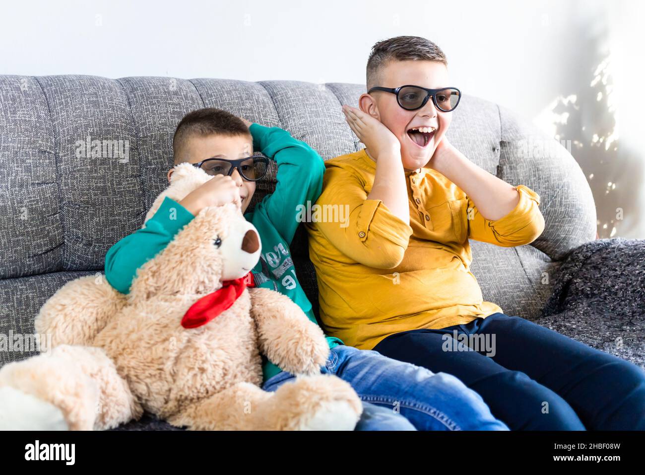 Cute boys, bothers watching movie with 3d glasses, sitting on the sofa in living room, together with teddy bear toy Stock Photo