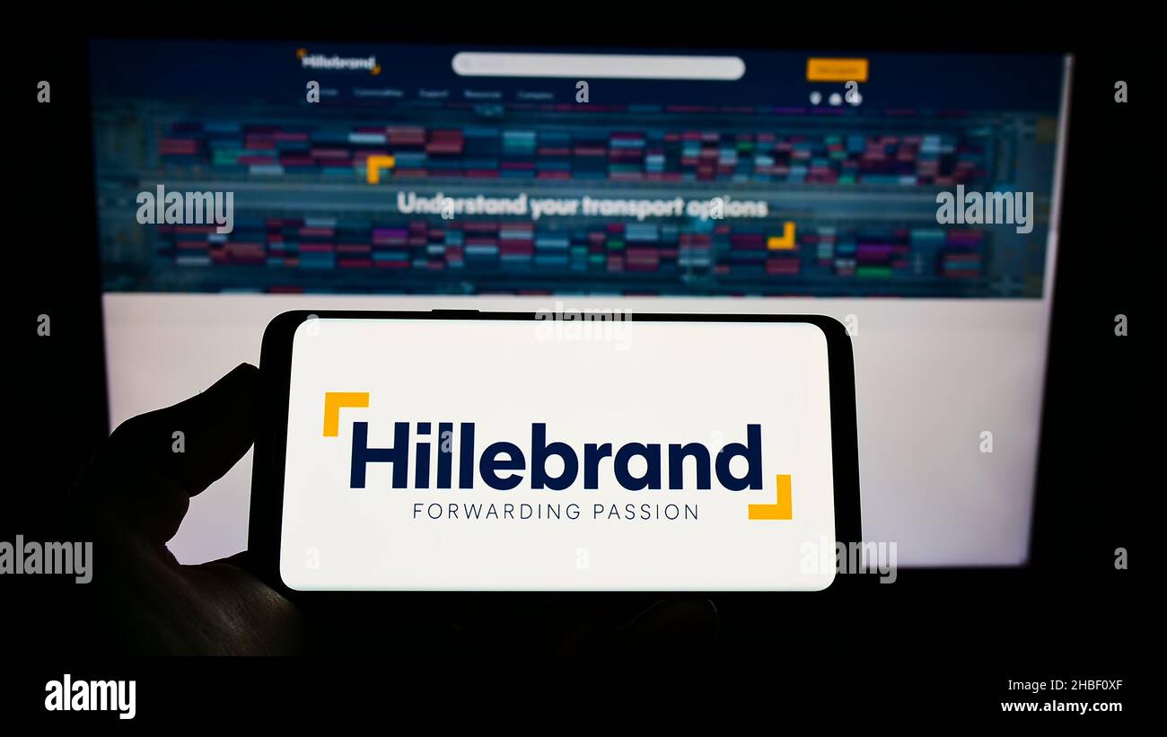 Person holding mobile phone with logo of German logistics company J.F. Hillebrand Group AG on screen in front of webpage. Focus on phone display. Stock Photo