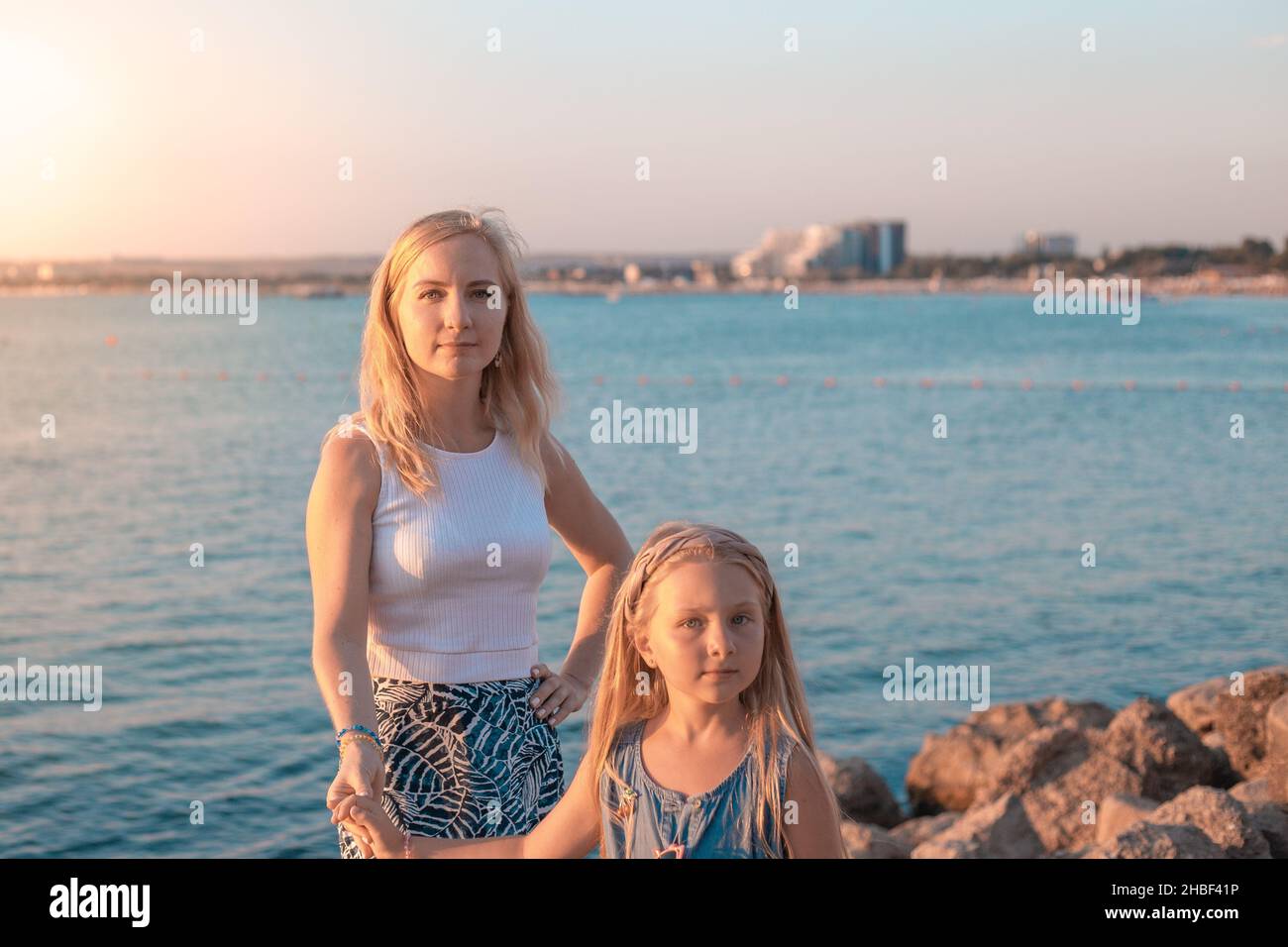 Two beautiful long-haired blondes mom and daughter on the seashore at sunset, hold hands, look at the camera. Stock Photo