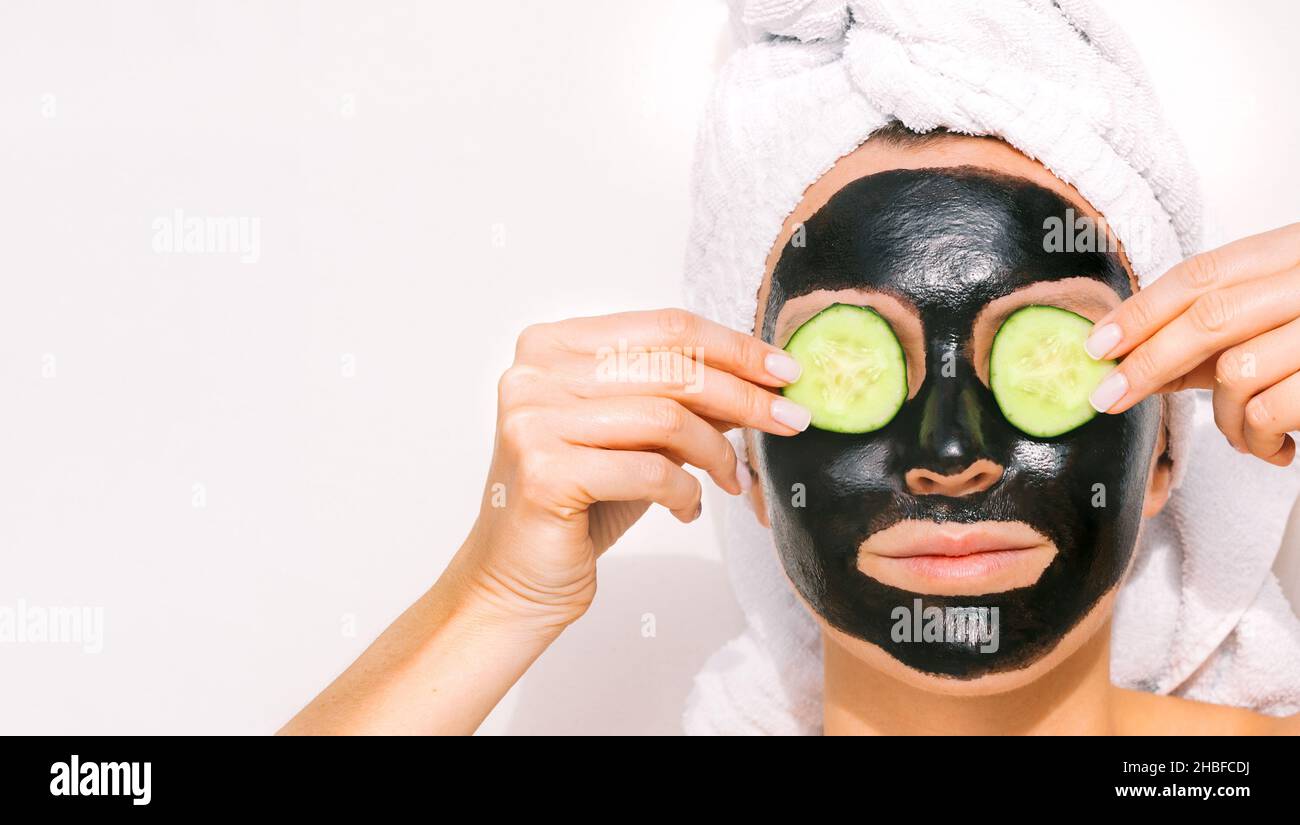 Woman applying black purifying charcoal facial mask with cucumber slices. Face skin care. Copy space Stock Photo