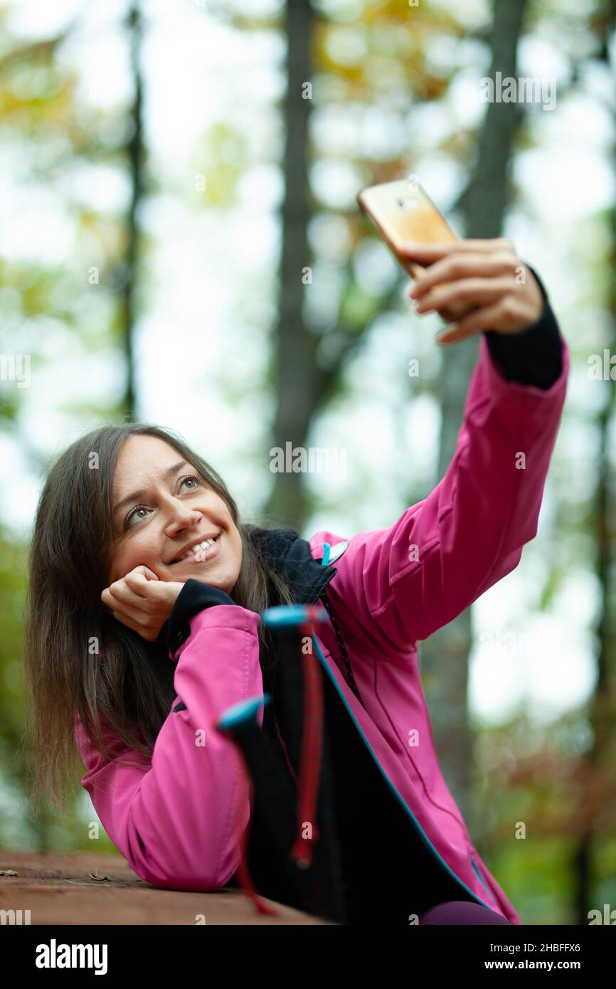 Hiker girl resting on a bench in the forest. Backpacker with pink jacket taking selfie with smartphone. Stock Photo