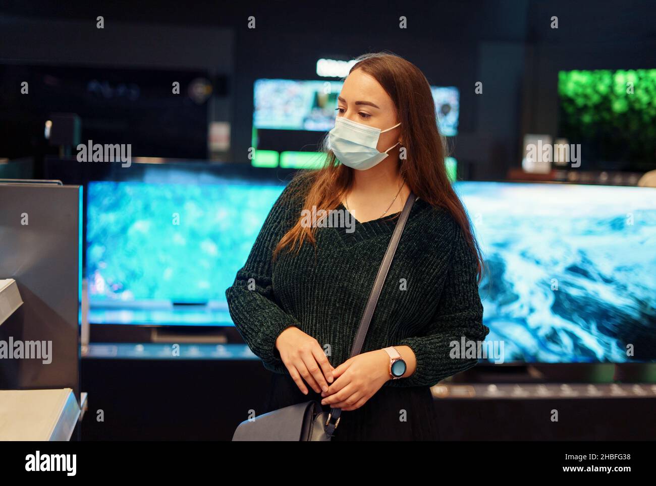 Young woman in medical face mask pensively examines variety of tv sets on display in tech store department during Covid19 outbreak, female doing shopping for home during pandemic Stock Photo