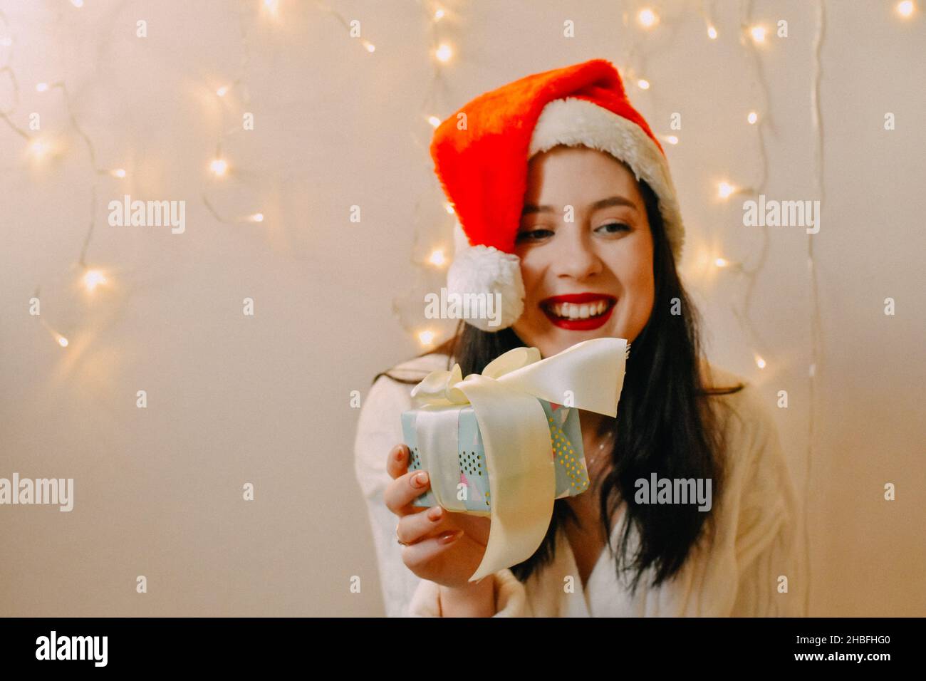 A closeup of a Caucasian girl wearing Santa hat and smiling with gift and Christmas lights on the wall Stock Photo