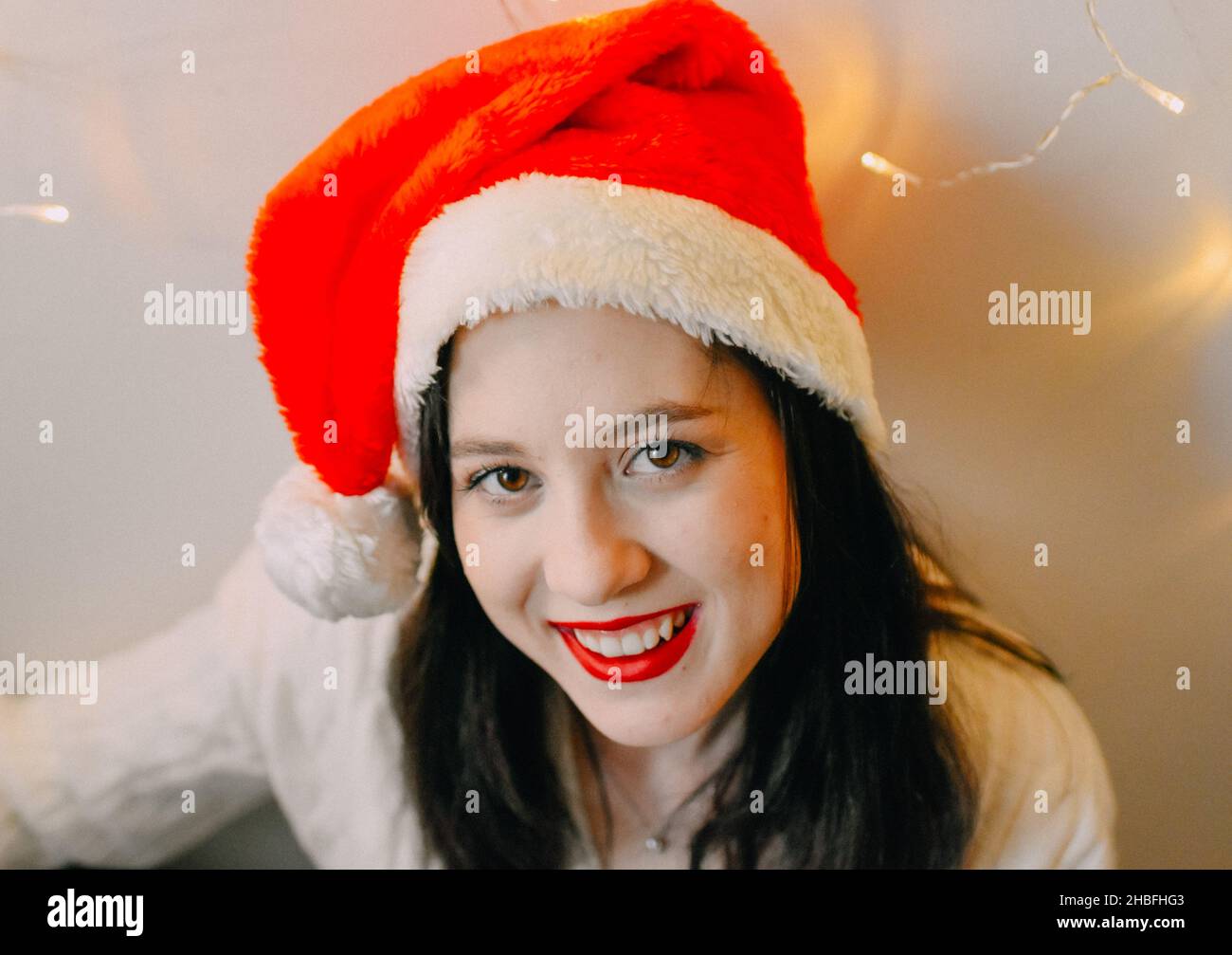 A closeup of a Caucasian girl wearing Santa hat and smiling with Christmas lights on the wall Stock Photo