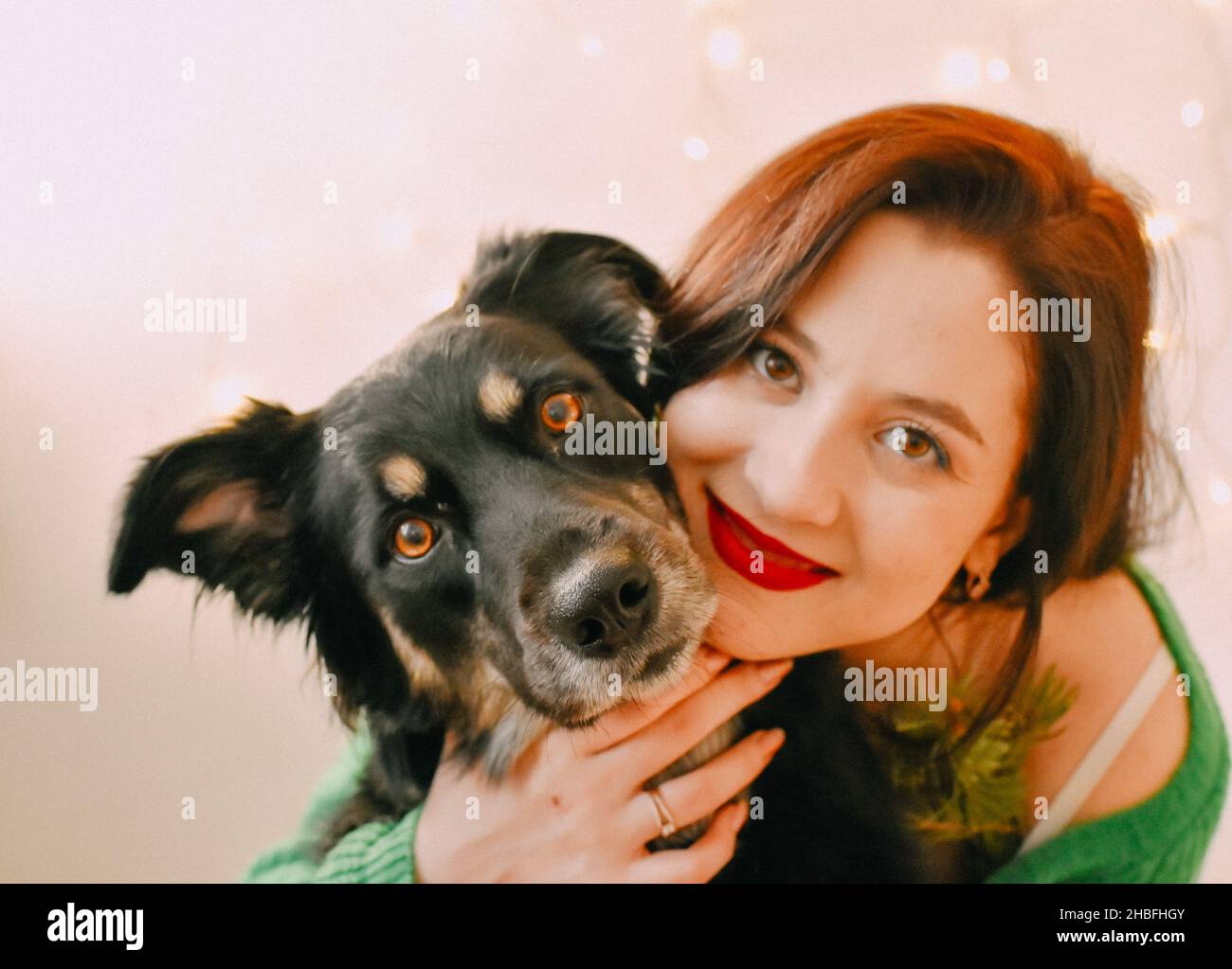 A closeup shot of a Caucasian girl smiling to the camera with her dog and Christmas lights on wall Stock Photo
