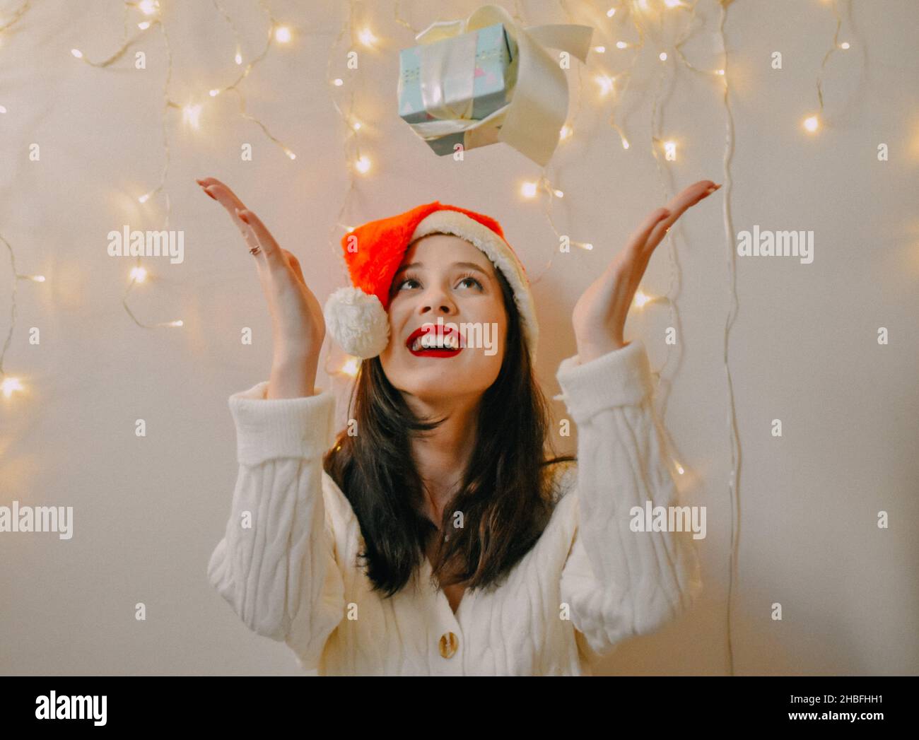 A shot of a Caucasian girl with Santa hat happy with her gift and Christmas lights on the wall Stock Photo