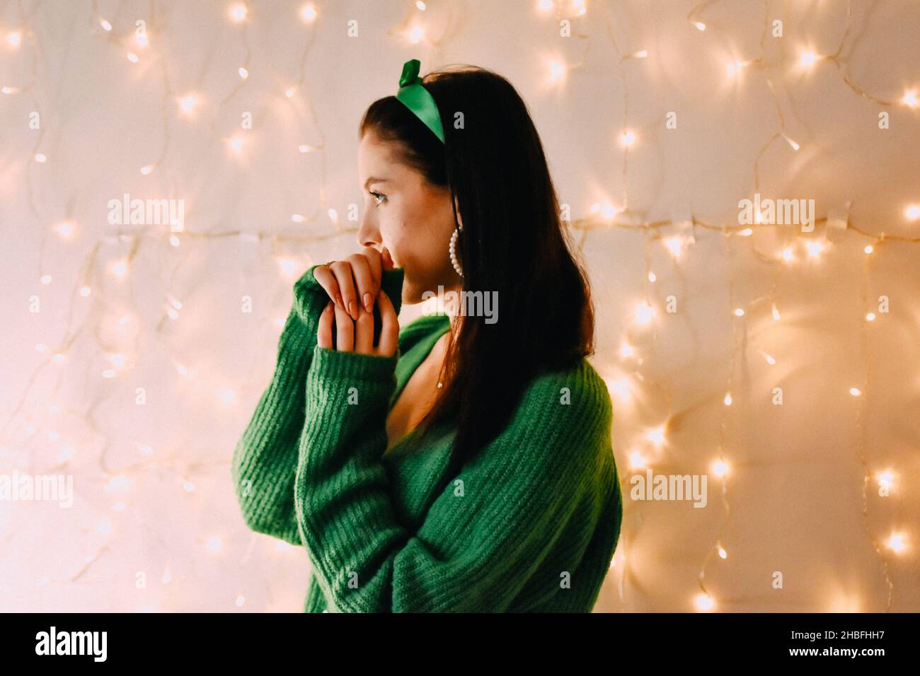 A closeup shot of the side profile of a Caucasian girl with green pullover and Christmas lights on wall Stock Photo