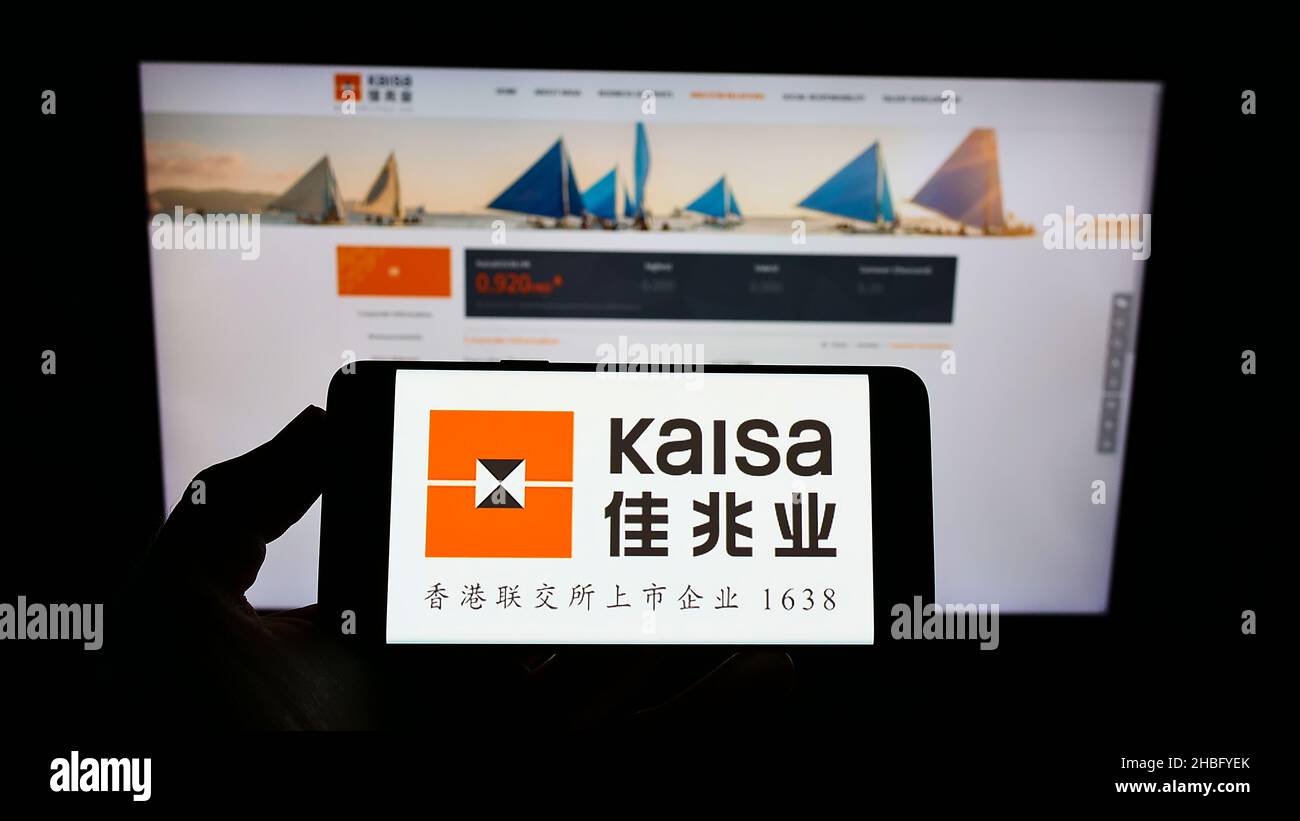 Person holding mobile phone with logo of Chinese property company Kaisa Group Holdings Ltd. on screen in front of web page. Focus on phone display. Stock Photo