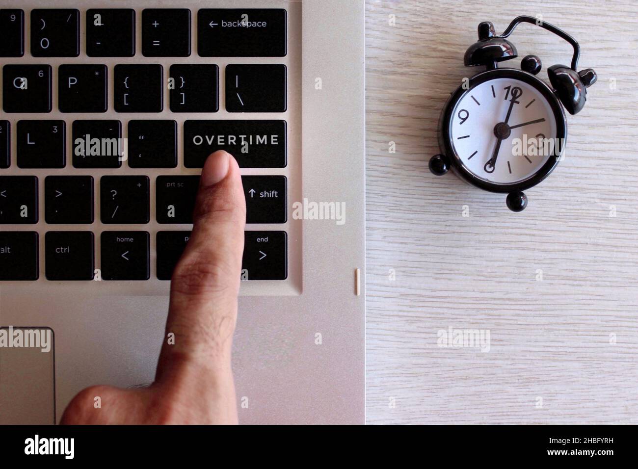 Overtime work concept. Finger pressing keyboard with text OVERTIME and black alarm clock. Stock Photo