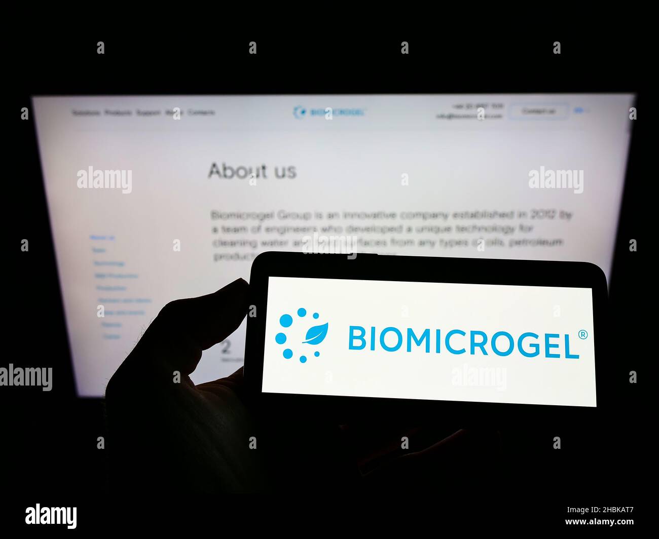 Person holding mobile phone with logo of British company Biomicrogels Group on screen in front of busines web page. Focus on phone display. Stock Photo