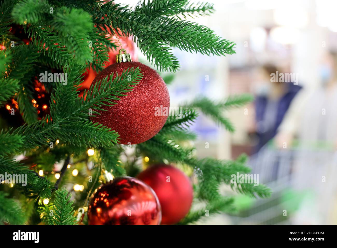 Christmas tree with red toy balls in a shopping mall on background of walking people in masks. New Year decorations, winter holidays Stock Photo