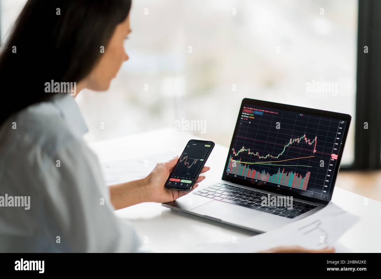 Young adult confident female investor, broker or crypto trader, using  laptop and smartphone, buys and sells crypto coins, analyzes charts of  trading in stock market and digital cryptocurrency exchange Stock Photo -