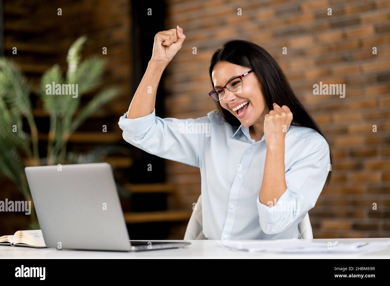 Successful cute confident emotional brunette caucasian broker, freelancer, business woman with glasses sitting at desk, using laptop, enjoying bargain, victory, good news, gesturing with fists, smily Stock Photo