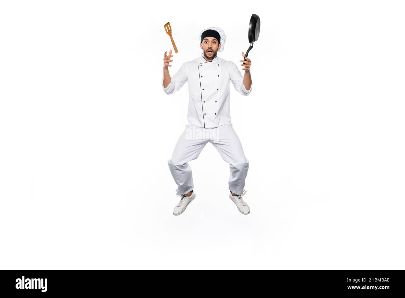 shocked chef in hat and uniform levitating near flying frying pan with wooden spatula isolated on white Stock Photo