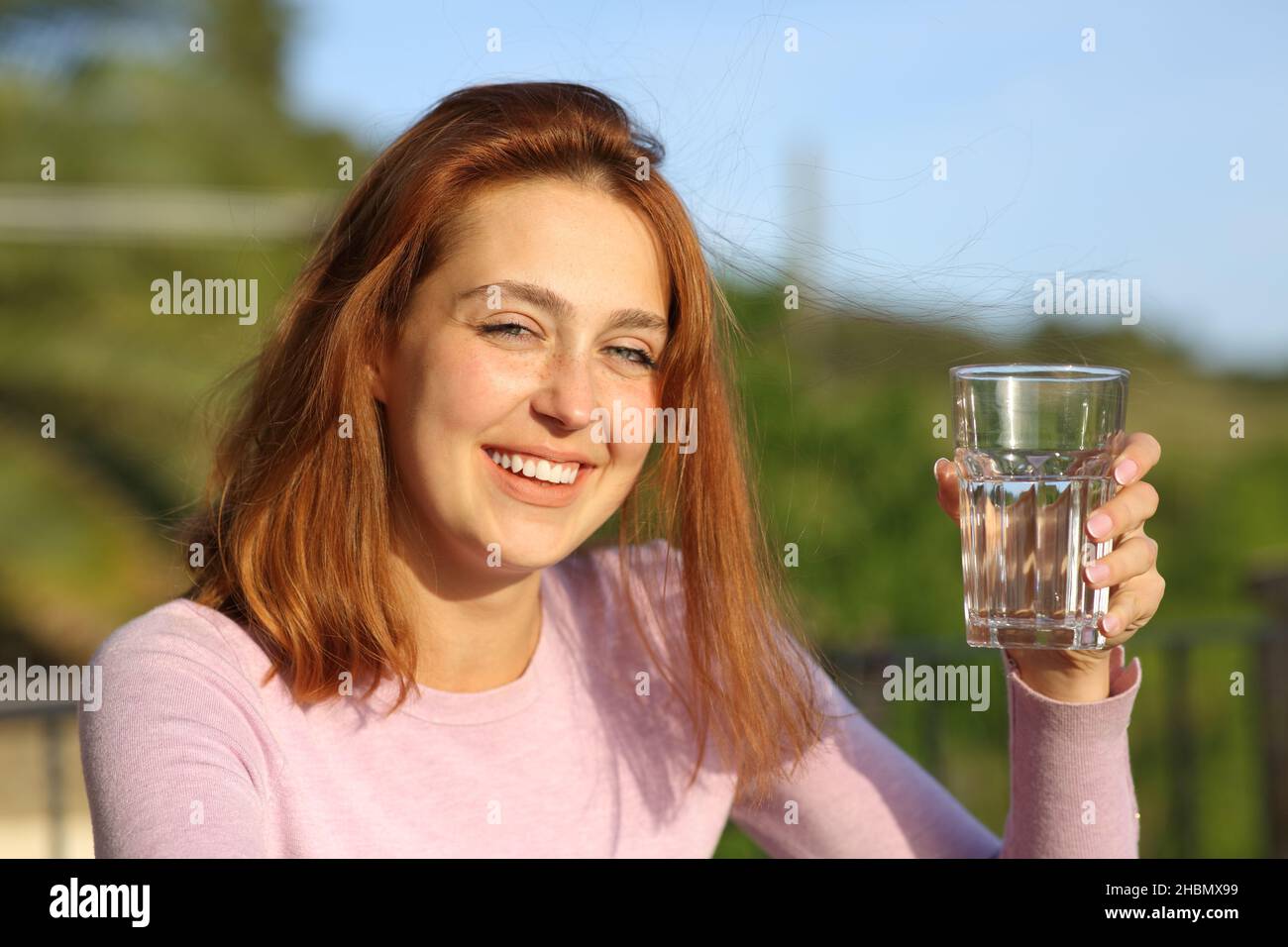 Happy woman holding glass of water smiling at camera in a balcony Stock Photo