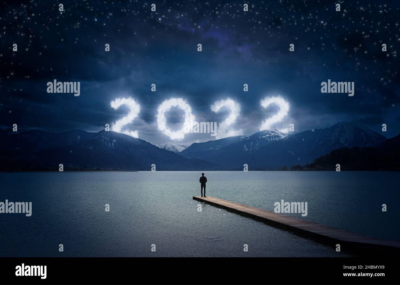 New year 2022 at night, man standing on a wooden dock on a lake and looking to the cloudy numbers in the dark sky over mountains, copy space Stock Photo
