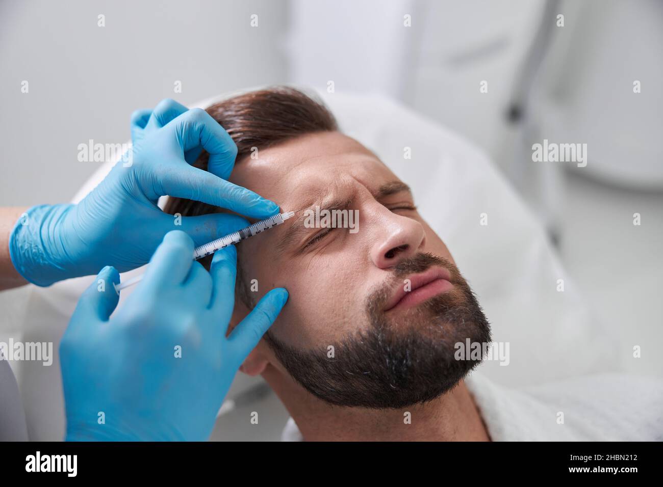 Ppatient grimaces of pain while careful cosmetologist does injection of lifting filler in his brow in clinic Stock Photo