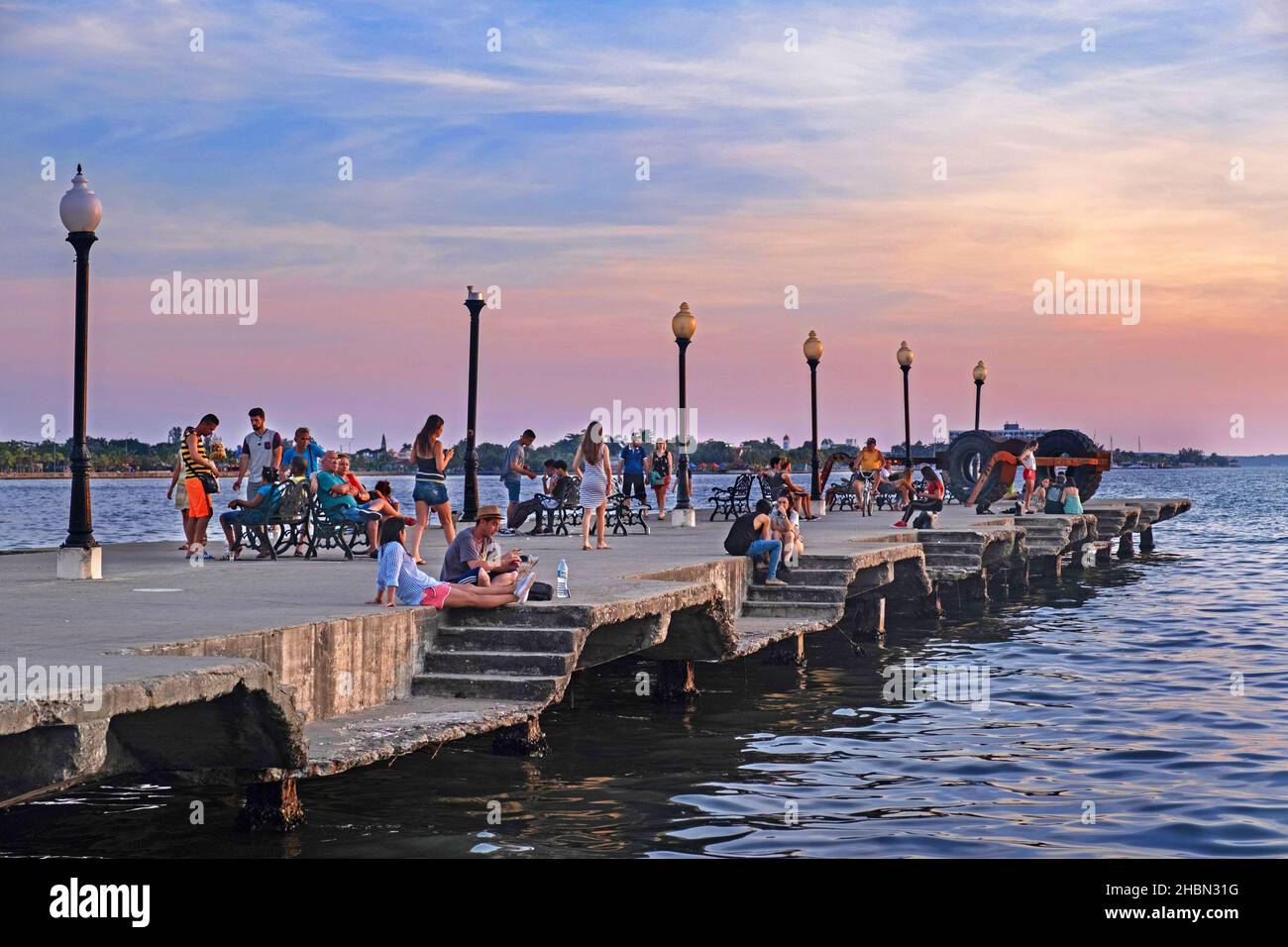 Cubans and tourists sitting on the concrete Muelle Real / Royal Pier at sunset in the city Cienfuegos on the island Cuba, Caribbean Sea Stock Photo