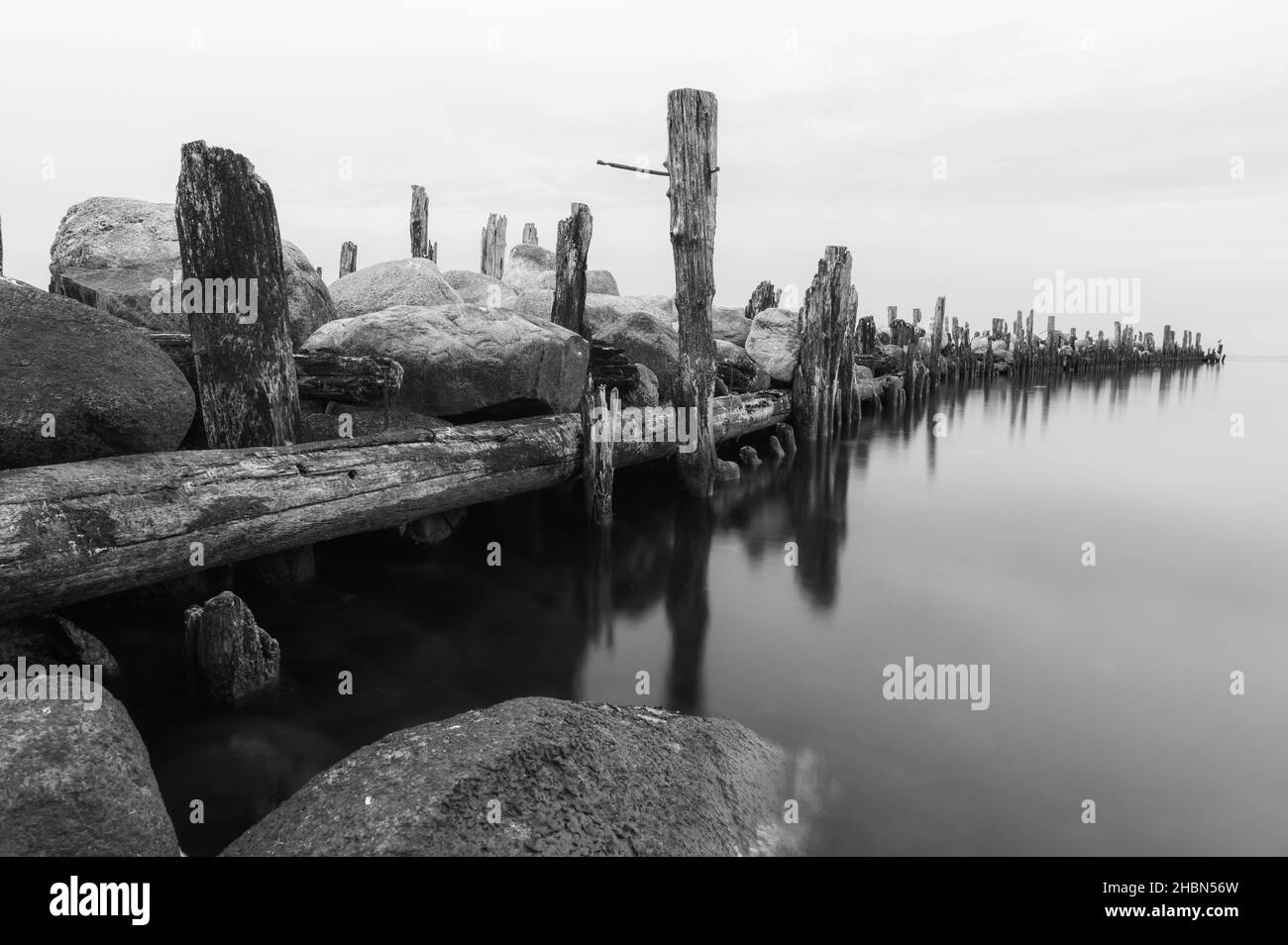 an old pier made of stones and wooden legs is left with metal screeds and the water surface is calm Stock Photo