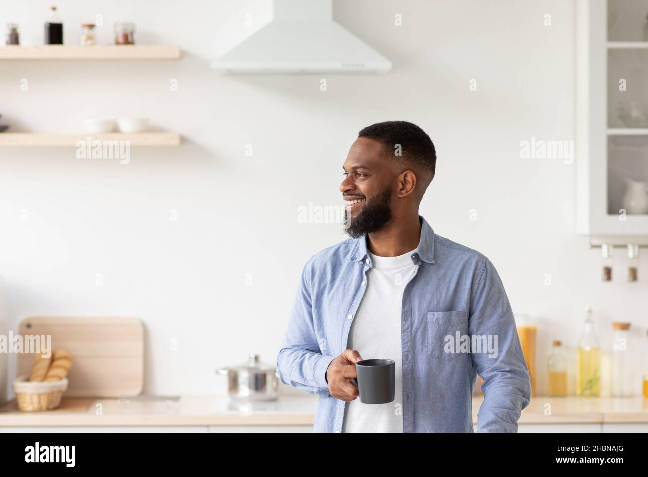 Happy young bearded african american guy with cup of coffee in hand in minimalist kitchen interior looks at empty space Stock Photo