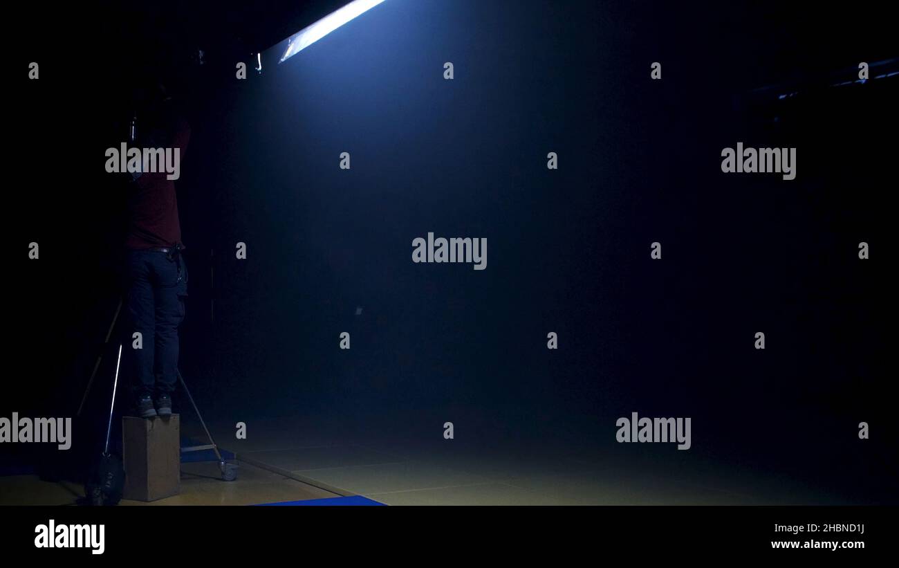 The film crew works on creation of a videoclip. Gaffer prepares diffusion for a light in the studio. Gaffer puts blue gel on a fresnel light on video Stock Photo