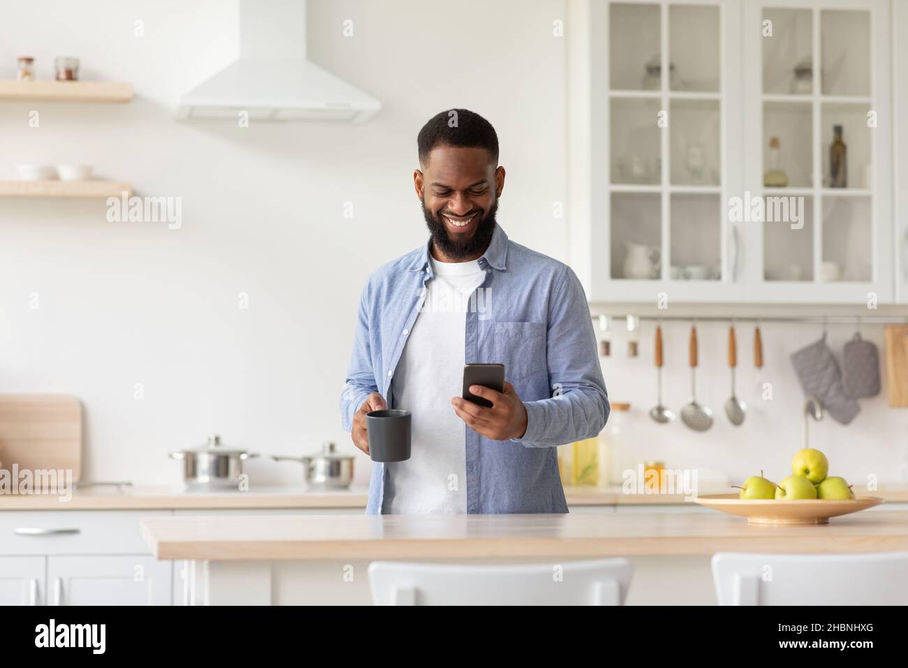 Cheerful young black male with cup of coffee in hand in minimalist kitchen interior reads news on smartphone Stock Photo